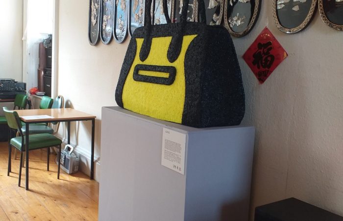 'Handbag' by artists Beagles & Ramsay is a sculpture of an oversized fluorescent yellow handbag, installed on a grey plinth at The Wing Hong Chinese Elderly Centre in Glasgow.