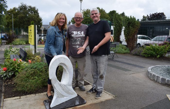 Three people gather around Tom Allan's sculpture 'Big Wave Vortex' which is installed in a small urban garden.