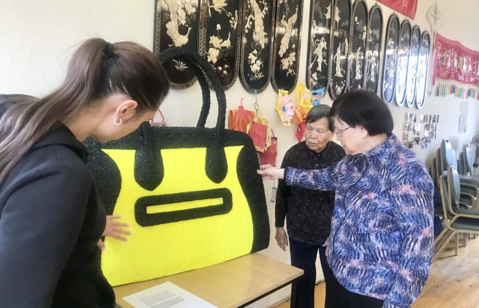 Three people view and touch the work 'Handbag', an oversized yellow handbag sculpture with black trim by Beagles & Ramsay, in Glasgow’s Wing Hong Chinese Elderly Centre.